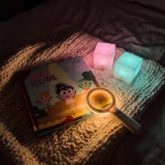 Lumi's First Dance Recital board book on a knitted blanket with the glowing Magic Magnifier and two light-up cubes
